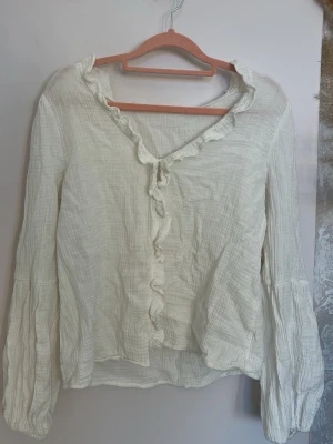 Vit volangblus i bomull - Söt vit blus i mjuk, krinklad bomull med volangkant längs v-ringningen och knäppning framtill. Långa ärmar med lätt puff och resår vid ärmslut. Perfekt för en romantisk och avslappnad stil.