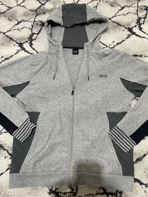 Gråmelerad hoodie från BOSS - Snygg gråmelerad hoodie från BOSS med dragkedja, huva och dragsko. Kontrastpaneler i mörkgrått på ärmar, fickor och huva samt randiga muddar vid handlederna. Tillverkad i mjuk bomullsblandning för extra komfort. Liten BOSS-logga broderad på bröstet.