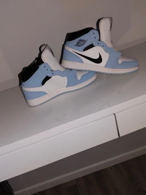 Jordan 1 High University Blue sneakers - Snygga Jordan 1 High sneakers i färgen University Blue med vita och svarta detaljer. Klassisk siluett med högt skaft, svart swoosh och blått Air Jordan-märke på sidan. Ovandel i läder och textil, perfekt för dig som gillar streetwear.