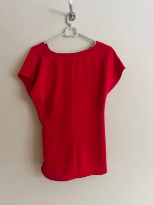 Röd knytblus från Zara - Snygg röd topp från Zara med korta ärmar och djup v-ringning framtill. Toppen har ett knytband i midjan som ger en schysst siluett och är gjord i ett mjukt, lätt material. Perfekt för dig som vill sticka ut med färg.