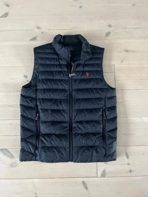 Mörkblå dunväst från Ralph Lauren - Snygg mörkblå dunväst från Ralph Lauren med diskret röd logga på bröstet. Västen har ståkrage, två dragkedjeförsedda fickor och quiltad design. Perfekt att slänga över en hoodie eller tröja för extra värme. Materialet är lätt och smidigt. Barnstorlek