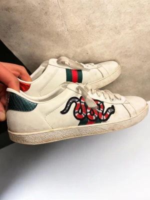 Gucci Ace sneakers  - Vita Gucci Ace sneakers i läder med broderad röd och svart orm på sidan samt klassiska gröna och röda ränder. Baksidan har metallicdetaljer i grönt och rött med Gucci-logga. Perfekta för dig som vill sticka ut med lyxig streetstyle.