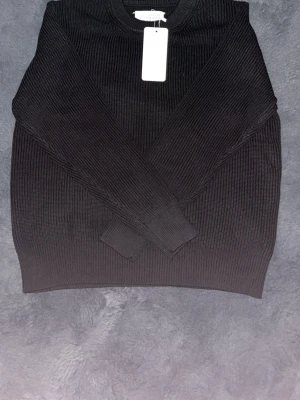 Svart stickad tröja Maison Margiela - Snygg svart stickad tröja från Maison Margiela med rund halsringning och ribbstickad struktur. Tröjan har långa ärmar och breda muddar vid ärmslut och nederkant. Perfekt för dig som gillar minimalistisk och stilren design.