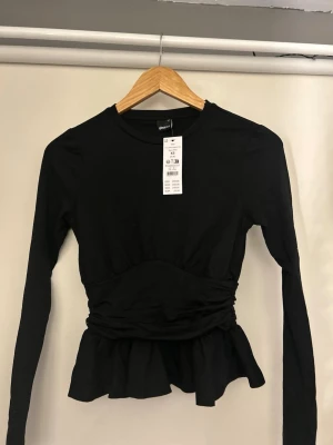 Svart långärmad topp med volang - Trendig svart långärmad topp från Gina Tricot med markerad midja och volang nedtill. Toppen har rund halsringning och är tillverkad i mjuk bomullsmix. Perfekt för dig som gillar en feminin och stilren look.