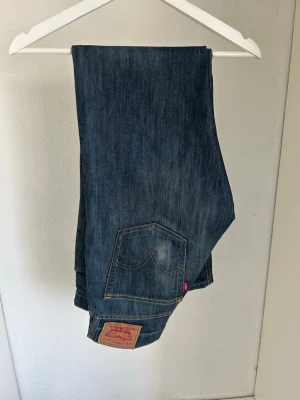 Levi's blå raka jeans - Klassiska blå jeans från Levi's i rak modell. Jeansen har fem fickor, synliga sömmar och den ikoniska läderlappen bak i midjan. Tillverkade i slitstarkt denim med en djupblå tvätt och diskret röd Levi's-tag på bakfickan. W32 l32