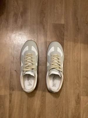 Maison Margiela gats vita - Stilrena sneakers i vitt och beige från maison margiela med klassisk retrodesign. Ovandelen är i en mix av mocka och syntet, med beige snörning och vit sula i gummi. Perfekta för en clean och avslappnad look.