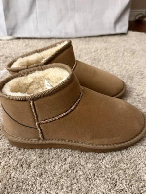 Bruna fluffiga tofflor från Nelly - Mysiga bruna tofflor från Nelly med mjukt, vitt foder på insidan. Tofflorna har en klassisk slip-on design med rund tå och är tillverkade i mockaimitation. Perfekta för att hålla fötterna varma hemma under kyliga dagar.