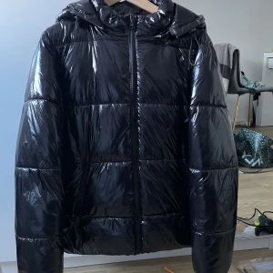 Svart glansig pufferjacka med huva - Säljer en svart, glansig pufferjacka med huva och dragkedja framtill. Jackan har en quiltad design med breda paneler och en oversized look. Perfekt för kalla dagar när du vill hålla dig varm och samtidigt se trendig ut. Storlek 36, nypris ca 1200kr