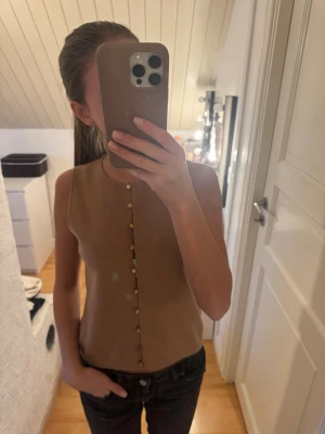 Beige ärmlös topp med knappar - Snygg beige ärmlös topp med rund halsringning och dekorativa knappar framtill. 
