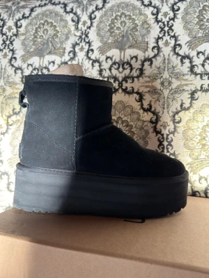 Svarta UGG moon boots med platåsula - Säljer ett par svarta UGG moon boots med hög platåsula. Boots i mocka med mjukt foder och klassisk UGG-detalj bak. Perfekta för kalla dagar och ger en cool look till vilken outfit som helst. Tidlös design och riktigt sköna att ha på sig.
