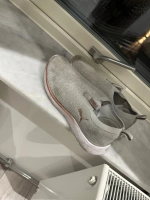 Grå slip-on sneakers från Puma - Säljer ett par grå slip-on sneakers från Puma med stickad ovandel och vit sula. Skorna har elastiska detaljer för enkel påtagning och en diskret Puma-logga. Den är använd. Storlek mellan 40-41