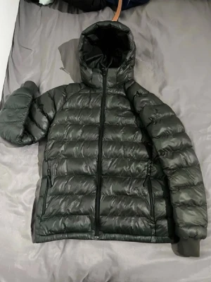 Svart dunjacka med huva - Snygg svart dunjacka med huva och dragkedja framtill. Jackan har två sidofickor med dragkedja och quiltad design som ger en puffig look. Perfekt för kalla vinterdagar och håller dig varm med sin vadderade insida.