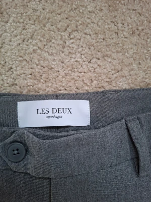 Grå kostymbyxor från Les Deux - Snygga grå kostymbyxor från Les Deux med klassisk rak passform och diskret knappstängning. Byxorna har en stilren look och är tillverkade i ett mjukt tyg som känns bekvämt hela dagen. Perfekta för dig som vill ha en clean och modern stil.