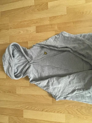 Grå hoodie från Lyle & Scott - Snygg grå hoodie från Lyle & Scott i 100% ekologisk bomull. Klassisk modell med huva och dragsko samt den ikoniska gula loggan på bröstet. Perfekt för en avslappnad och stilren look.