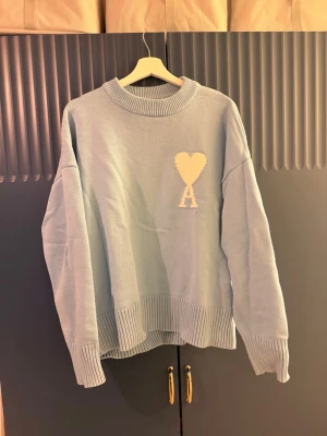 Ljusblå stickad tröja Ami Paris - Ljusblå oversized stickad tröja från Ami Paris med ribbade muddar och nederkant. Tröjan har ett vitt hjärta och bokstaven 'A' broderat på bröstet. Rund halsringning och avslappnad passform, perfekt för en chill look.