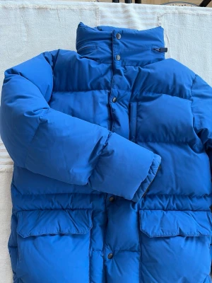 Dunjacka Aspesi - Dunjacka från det italienska lyxmärket Aspesi. Jackan är i bra skick med några få tecken på användning (främst  på dragkedjan, se bild). Färgen är en mycket fin klarblå nyans och fodret är svart med orange detaljer.  Storlek Medium, men den är liten i storleken och passar därför troligen någon som normalt bär Small.  Många fina detaljer, som  huva som fälls in osynligt i kragen, snölås i midjan samt två innerfickor.  Säljes pga köpt fel storlek.  Nypris ca 7000 SEK