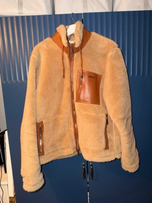Axel Arigato Fleece jacka - Säljer en beige teddyjacka från Axel Arigato med detaljer i brunt konstläder på krage, fickor och axel. Jackan har dragkedja framtill, hög krage med snörning och tre fickor med dragkedja. Perfekt för dig som vill ha en mysig och trendig look.