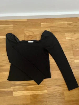 Svart långärmad topp med puffärm - Trendig svart topp med lång ärm och puffiga axlar. Toppen har en croppad passform och är tillverkad i ett stretchigt, smockat material som ger en snygg struktur. Perfekt för dig som vill ha en stilren men ändå unik look.