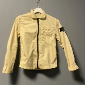 Gul jacka från Stone Island - Säljer en ljusgul jacka från Stone Island med klassisk patch på vänster ärm. Jackan har dragkedja framtill, bröstficka och krage. Perfekt för dig som gillar streetwear och vill sticka ut med en unik färg. Ny pris 3500 