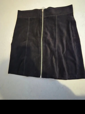 Svart manchesterkjol med dragkedja - Säljer en svart kortkjol i manchester med en snygg silverfärgad dragkedja framtill. Kjolen har en rak passform och är perfekt för dig som gillar en enkel men trendig stil. Passar till många olika looks och är superenkel att matcha.