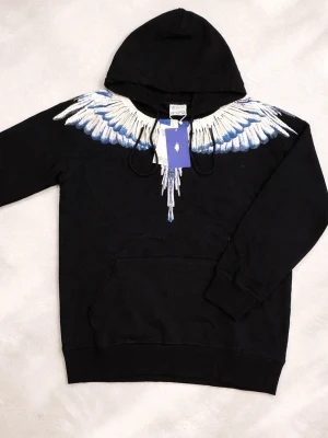 Marcelo Burlon hoodie - Säljer en svart hoodie från Marcelo Burlon med ikoniskt vingtryck i vitt och blått över axlar och bröst. Hoodien har huva med dragsko och en stor magficka. Perfekt för dig som vill sticka ut med streetwear-vibe. Helt ny!!