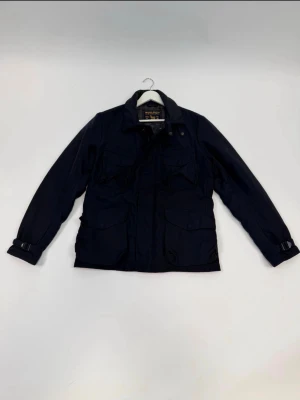 Svart vindjacka från woolrich - Stilren svart vindjacka från woolrich med fyra framfickor och justerbara spännen vid ärmsluten. Jackan har klassisk krage och stängs med dragkedja och knappar. Perfekt för dig som gillar en tidlös look. Storlek Xl/L men passar L bäst 