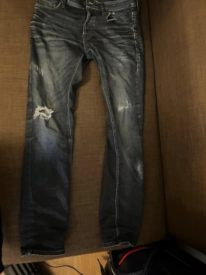 Slim fit jeans från Jack & Jones - Säljer ett par mörkblå slim fit jeans från Jack & Jones, modell Glenn. Jeansen har slitna detaljer och hål på ena benet för en trendig, avslappnad look. Klassisk femficksmodell med dragkedja och knapp. Perfekta för dig som gillar streetstyle.