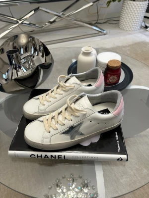 Golden Goose sneakers med rosa detalj - Snygga sneakers från Golden Goose i vitt läder med grå mockadetaljer och rosa häl. Klassisk stjärna i silver på sidan och beigea skosnören. Perfekta för dig som gillar en clean men ändå trendig look.