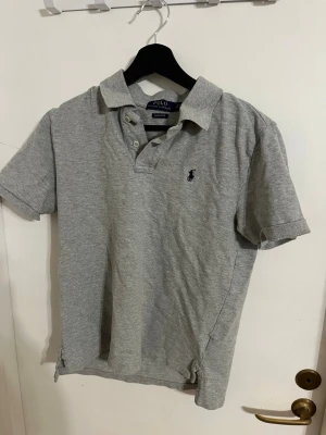 Grå pikétröja från Polo Ralph Lauren - Klassisk grå pikétröja från Polo Ralph Lauren med korta ärmar och krage. Tröjan har knappar vid halsen och den ikoniska broderade loggan på bröstet. Tillverkad i  bomull för en bekväm känsla.