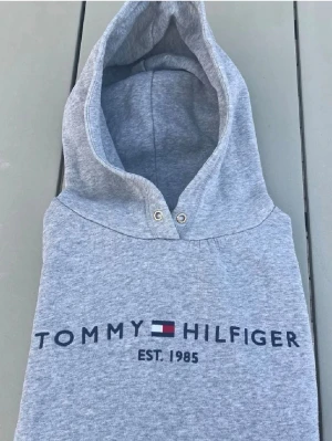 Grå hoodie från Tommy Hilfiger - Snygg grå hoodie från Tommy Hilfiger med klassisk logga och texten 'EST. 1985' framtill. Hoodien har huva med metallöljetter och är tillverkad i mjuk bomull. Perfekt för en avslappnad och stilren look.