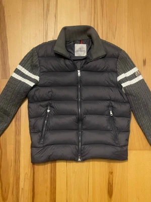 Grå dunväst från Moncler - Snygg grå dunväst från Moncler med stickade ärmar och vita ränder på överarmarna. Västen har hög krage, två dragkedjeförsedda fickor och quiltad front. Perfekt för dig som vill ha en sportig och trendig look.