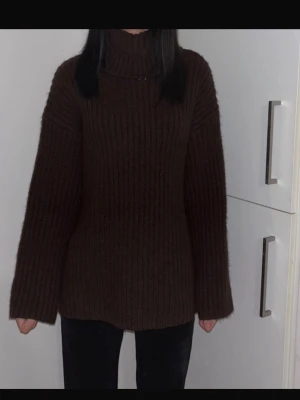 Brun ribbstickad polotröja - Mysig brun stickad polotröja med breda ribbor och långa ärmar. Tröjan har en hög polokrage och är tillverkad i ett tjockare garn som ger en avslappnad och trendig look. Perfekt för kalla dagar och enkel att styla med jeans eller kjol.