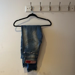 Blå slitna jeans från Dsquared2 - Säljer ett par blå jeans från Dsquared2 med kraftiga slitningar och hål på benen. Jeansen har en klassisk femficksmodell, låg midja och smal passform. Detaljerad med en röd Dsquared2-lapp bak och slitna kanter för en cool, trendig look.