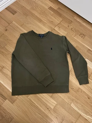Polo Ralph Lauren - Snygg olivgrön stickad tröja från Polo Ralph Lauren med klassisk rund halsringning och den ikoniska mörkblå logotypen broderad på bröstet. Tröjan har långa ärmar och ribbade muddar vid ärmslut och nederkant. Perfekt för en avslappnad men stilren look. Size: M 10-12 år