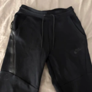Svarta nike tech mjukisbyxor  - Snygga svarta mjukisbyxor från Nike med dragsko i midjan och diskret logga på benet. Perfekta för en avslappnad stil eller träning. Byxorna har praktiska fickor med dragkedja.