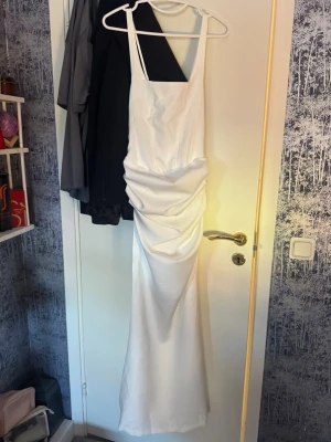 Vit långklänning med snörning - Elegant vit långklänning med smala axelband och snygg drapering framtill. Klänningen har snörning i ryggen för justerbar passform och är tillverkad i ett glansigt material som ger en lyxig känsla. Perfekt för speciella tillfällen. Dock var den för liten när jag fick hem den köpt för 1200kr
