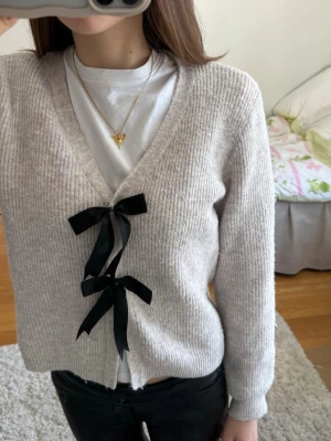 Beige kofta med svarta rosetter H&M - Supersöt beige ribbad kofta från H&M med två svarta rosetter framtill. Koftan har v-ringning, långa ärmar och mjukt stickat material som känns riktigt mysigt. 