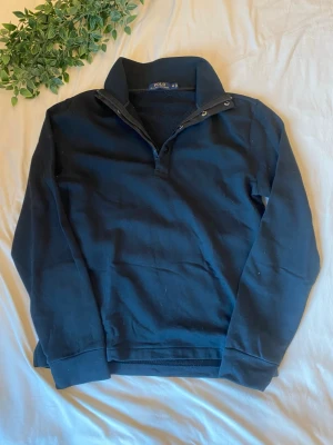 Svart tröja från Polo Ralph Lauren - Svart långärmad tröja från Polo Ralph Lauren med hög krage och knappslå. Tröjan har ribbade muddar vid ärmslut och nederkant, samt en klassisk och stilren design som passar till det mesta.