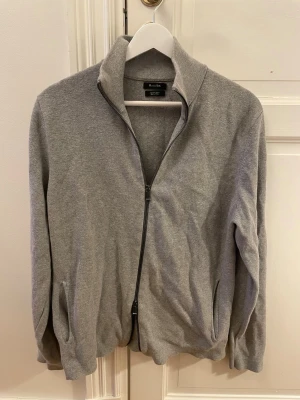Grå ziptröja från Massimo Dutti - Stilren grå ziptröja från Massimo Dutti med hög krage och hel dragkedja framtill. Tröjan har två fickor på sidorna och ribbade muddar vid ärmslut och nederkant. Perfekt för lager på lager och enkel att matcha.