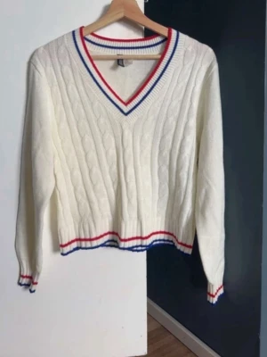 Vit kabelstickad tröja med ränder - Vit kabelstickad tröja med V-ringning och ribbade muddar. Röd och blå randdetalj vid hals, ärmslut och nederkant ger en lite elegant vibe. Croppad modell som passar perfekt till jeans eller kjol. Mjukt material. Använt en gång!💕☀️
