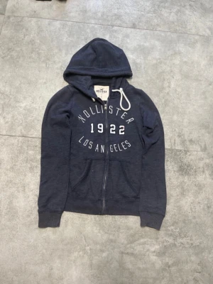 Mörkblå Hollister hoodie med dragkedja - Mörkblå hoodie från Hollister med vit text 'Hollister 1922 Los Angeles' på bröstet. Tröjan har huva med vita snören, dragkedja framtill och två fickor. Perfekt för en avslappnad och cool stil.
