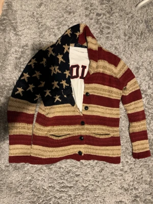 Stickad kofta med USA-flagga - Snygg stickad kofta med USA-flagga, röd, beige och mörkblå med stjärnor och ränder. Koftan har knappar framtill, sjalkrage och långa ärmar. Perfekt för dig som vill sticka ut med en unik och färgstark look. Cardigan från Ralph Lauren. 