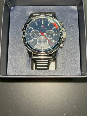 Blå klocka från Tommy Hilfiger - Snygg blå klocka från Tommy Hilfiger med rund urtavla, silverfärgade detaljer och svart länkarmband i metall. Urtavlan har flera små visare och röda detaljer som ger en sportig känsla. Perfekt accessoar för dig som gillar stilrena klockor. Köpte klockan för ungefär 2 995 ny. 