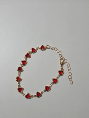 Guldarmband med röda hjärtan - Snyggt armband i guldfärgad metall med små röda hjärtan längs hela kedjan. Hjärtana har en glansig finish och armbandet har justerbar längd med karbinlås. Perfekt accessoar för att addera lite färg och kärlek till din stil.                     Detaljer: Färg: Guldton med klara röda hjärtan Huvudmaterial: Legering  Sten: Artificiell kristall (röd) Design: Länkarmband med hjärtformade stenar Längd: Ca 18 cm + 7 cm förlängningskedja Mått: 6 cm (i cirkel) Skick: Helt nytt / Oanvänt