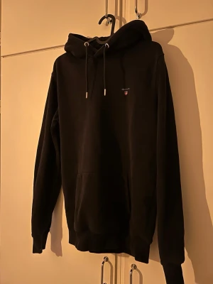 Svart hoodie från Gant med logga - Svart hoodie från Gant med liten broderad logga på bröstet. Klassisk känguruficka framtill och justerbar huva med snören. Mjuk bomullsblandning och ribbade muddar vid ärmslut och nederkant. Perfekt för en chill och stilren look.