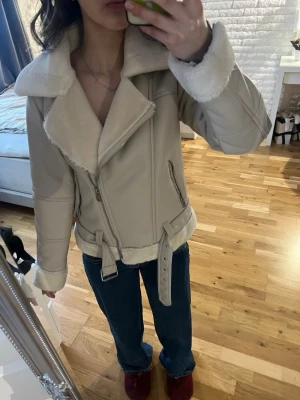 Beige pälsjacka med bälte - Snygg beige pälsjacka med bred krage och mjukt foder. Jackan har dragkedja, två fickor med dragkedja och ett bälte i midjan. Pälsdetaljer vid krage, ärmslut och nederkant ger en lyxig känsla. Perfekt för kalla dagar och trendig stil.