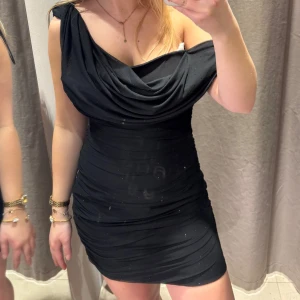 Svart kort klänning med drapering - Snygg svart kort klänning med draperad urringning och smal passform köpt ifrån Zara. Aldrig använd med prislapp kvar. Materialet känns stretchigt och följsamt. Glittrig och svart