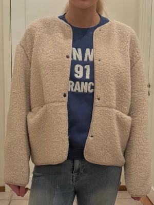 Beige teddyjacka med fickor - Mysig beige teddyjacka med rund halsringning och stora framfickor. Jackan har tryckknappar framtill och är gjord i fluffigt teddy-material som ger en avslappnad och trendig look. Perfekt för att hålla värmen under kyliga dagar.