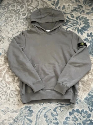 Stone Island hoodie - Snygg grå hoodie från Stone Island med klassisk patch på vänster ärm. Hoodien har en stor känguruficka framtill och ribbade muddar vid ärmslut och nederkant. Tillverkad i mjuk bomull för skön känsla och avslappnad stil.