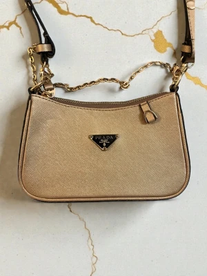 Beige axelremsväska från P - Stilren beige handväska från Pa i saffiano-läder med guldfärgade metalldetaljer och kedja. Väskan har klassisk rektangulär form, dragkedja upptill och Pas ikoniska logga framtill. Perfekt för dig som gillar lyxiga accessoarer och trendig design.